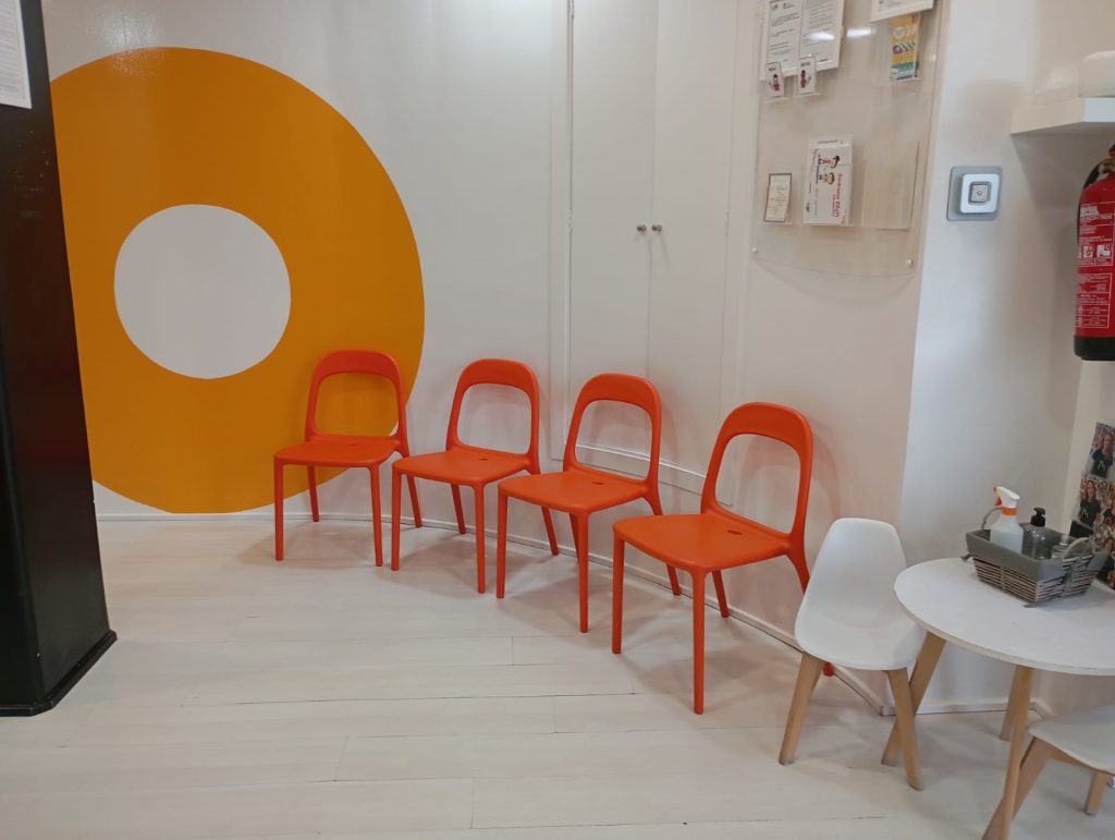 Sala de espera minimalista con sillas rojas y una mesa blanca sobre un suelo de madera clara, junto a una pared con un gran círculo naranja.