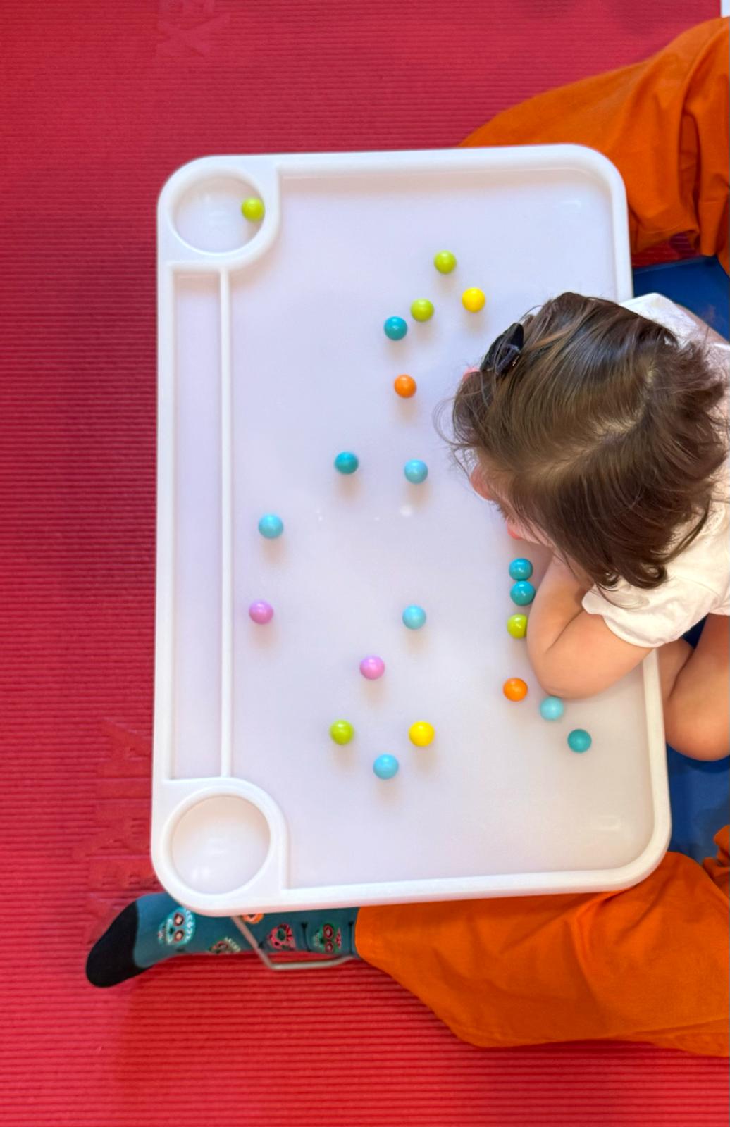 Niño concentrado en recoger bolas de colores de una bandeja blanca sobre una colchoneta roja.