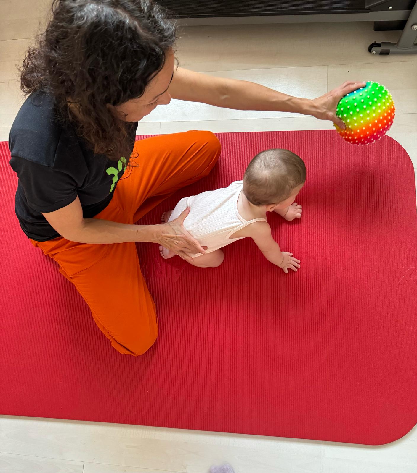 Adulto interactuando con un bebé en una colchoneta de ejercicios roja, ofreciéndole una pelota de colores.