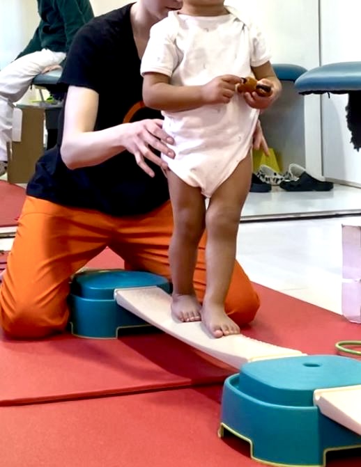 Un niño de pie sobre una tabla de equilibrio recibe apoyo y guía de un adulto que se encuentra arrodillado a su lado.