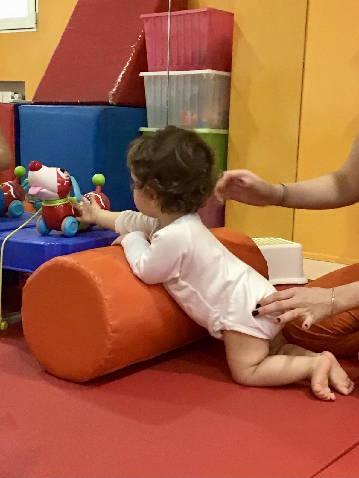 Niño pequeño en camiseta blanca jugando con un juguete sobre un rollo de espuma naranja, con la mano de un adulto en su espalda.